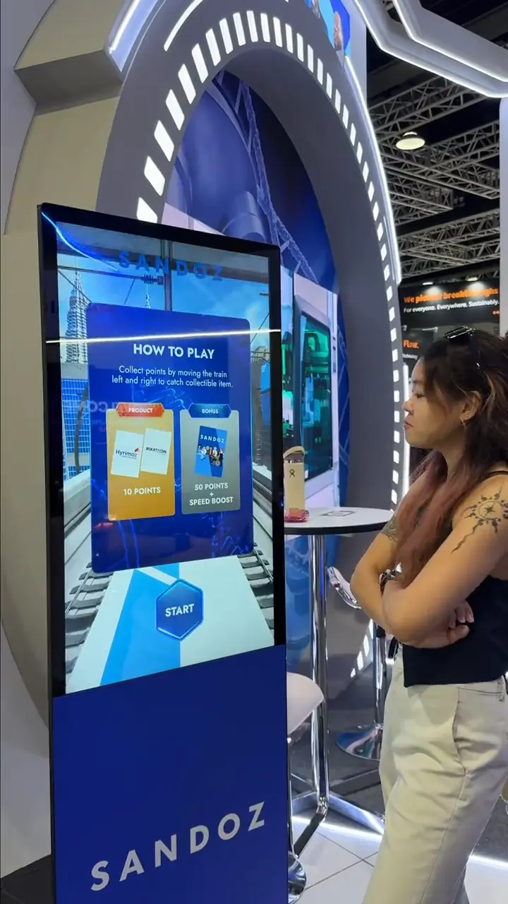 Sandoz game kiosk showing the "How to Play" instructions screen as a visitor looks on at the exhibition stand