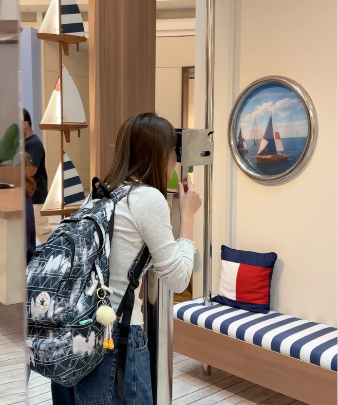 A visitor with a backpack peers into the viewfinder beside a porthole picture of sailboats