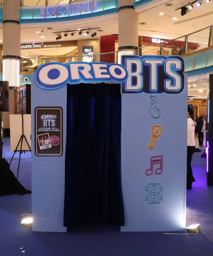 Front view of the light blue Oreo BTS photobooth in the mall's Blue Atrium, with a navy curtain entrance and neon K-pop icons of a finger heart, light stick, music note, and speaker on the side panel