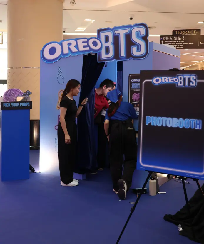 Visitors entering the Oreo BTS photobooth through the navy curtain while a staff member in a blue uniform assists, beside the glowing "Photobooth" easel sign