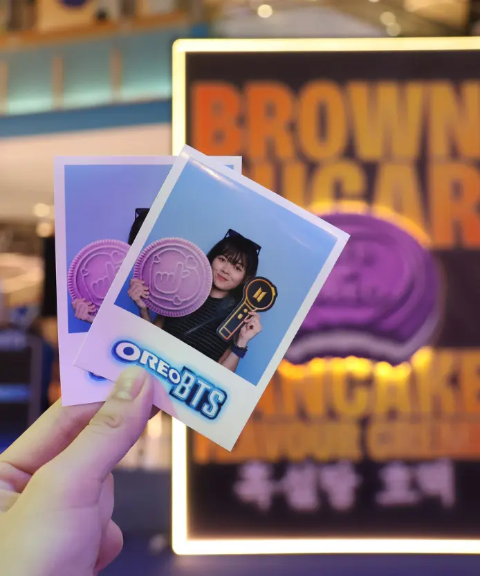 Hand holding two printed Oreo BTS instant photos of a visitor posing with a purple cookie prop and BTS light stick prop, in front of a Brown Sugar flavour display
