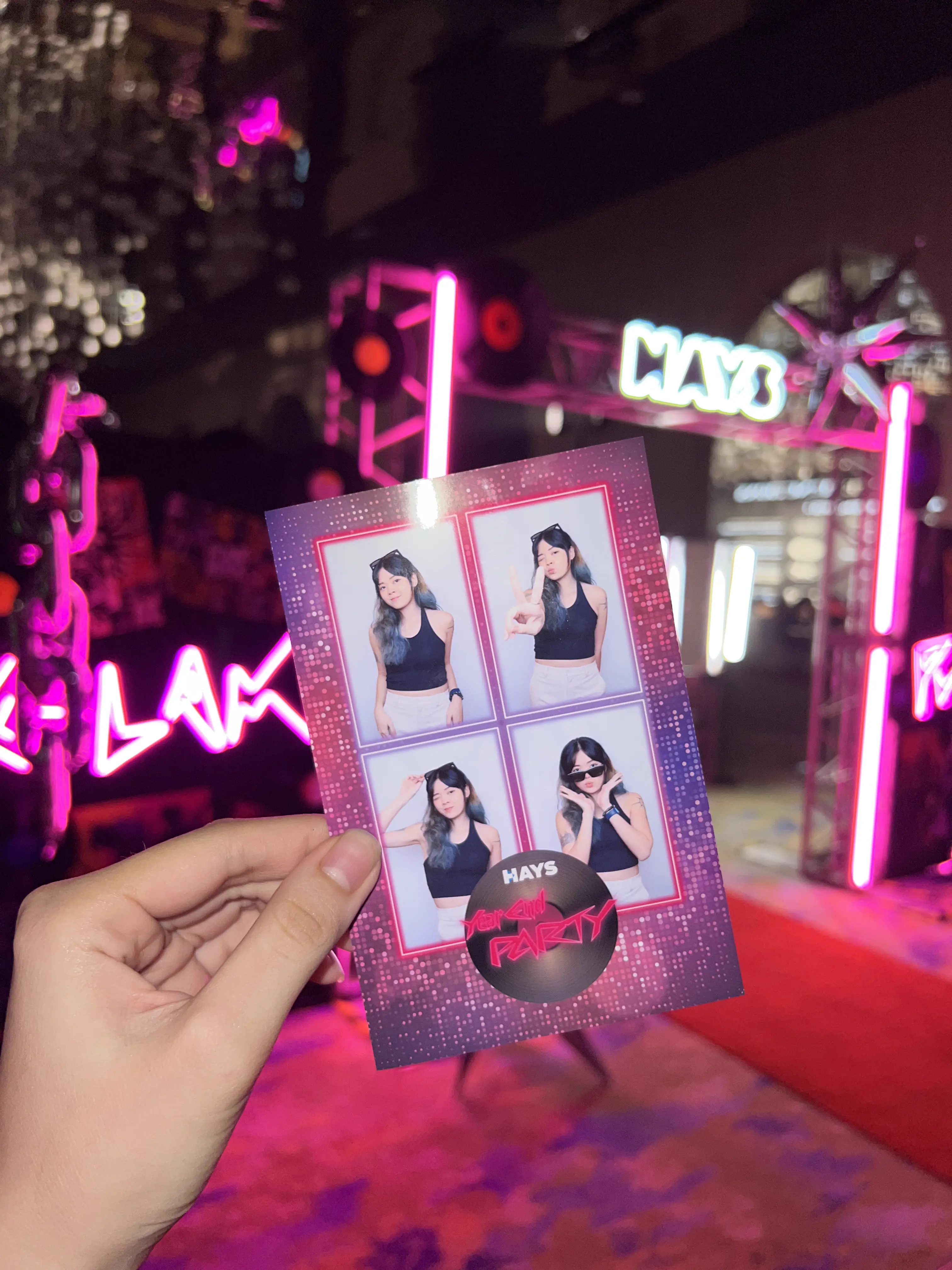 Hand holding a HAYS Year End Party photo strip in front of the neon arch