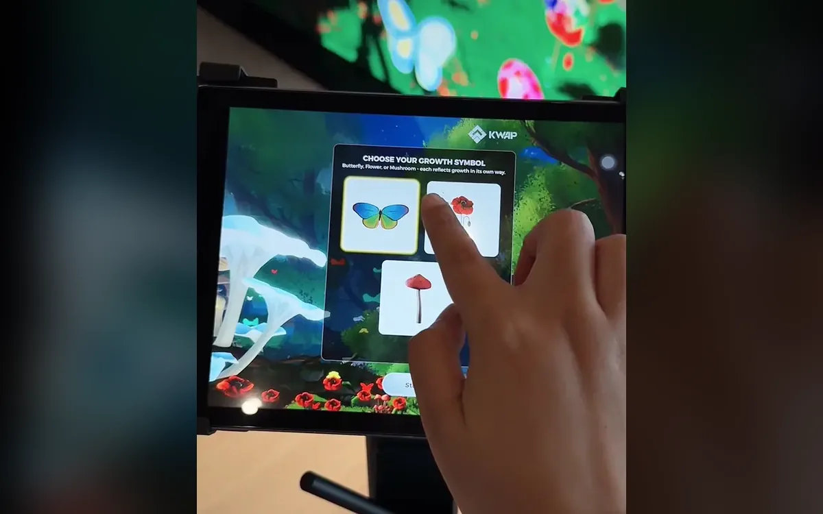 A hand taps a growth symbol, butterfly, flower or mushroom, on a KWAP iPad station with a garden scene glowing on the screen above