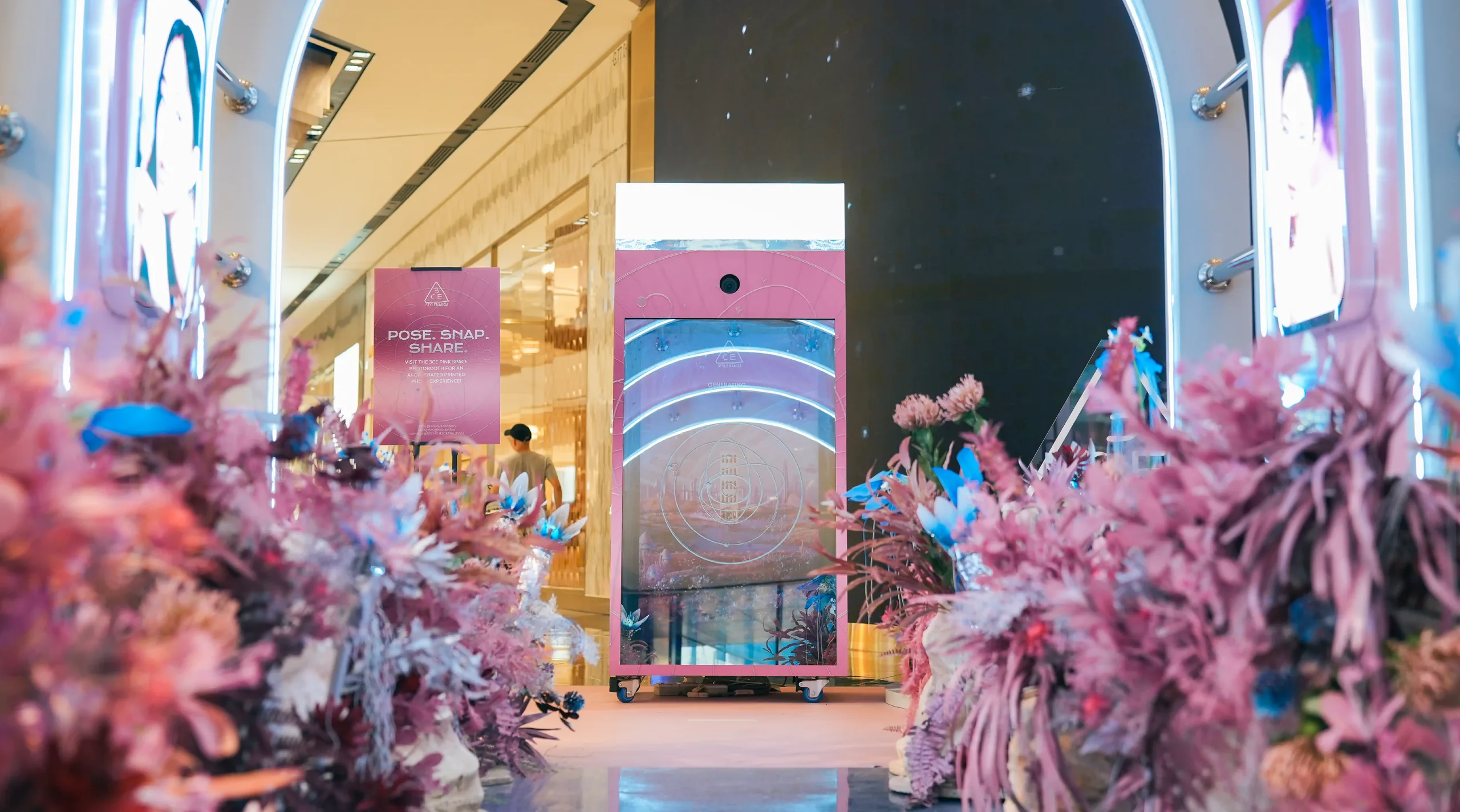 A pink 'Pose, Snap, Share' photo booth kiosk set among decorative floral displays in a mall.
