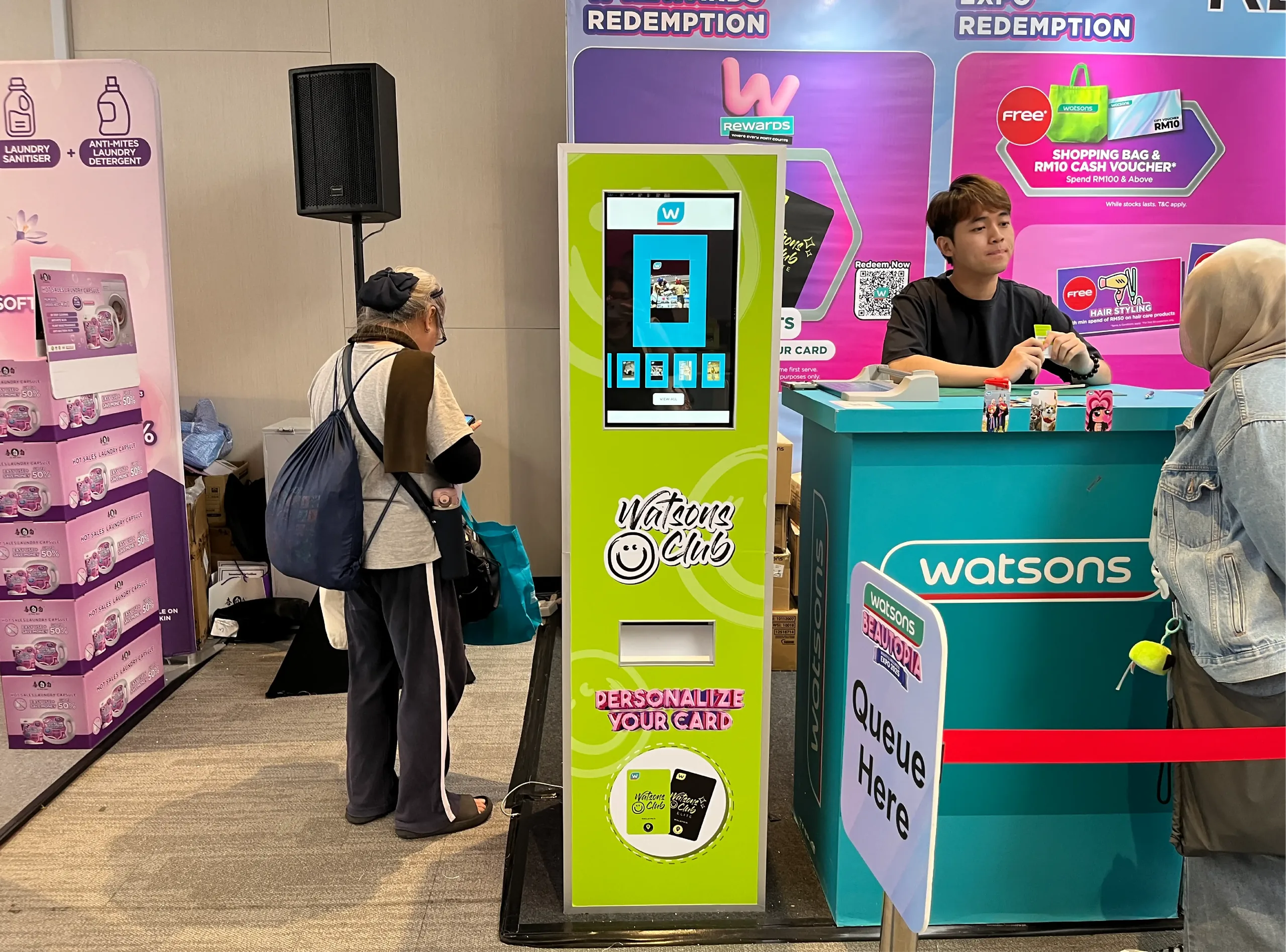 A green membership card kiosk stands beside a staffed redemption counter at a retail expo.