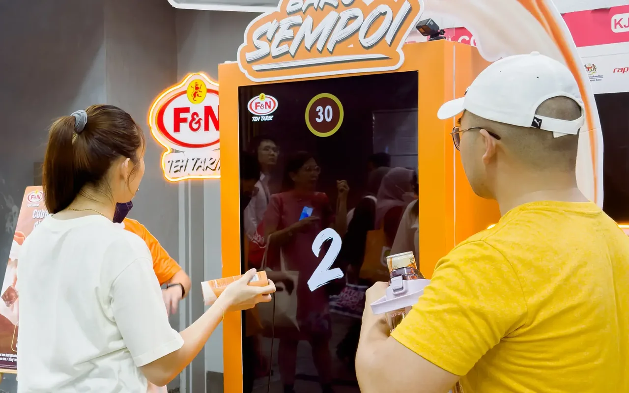 Dua pengunjung bermain cabaran mesin layan diri goncang-untuk-menang F&N Teh Tarik