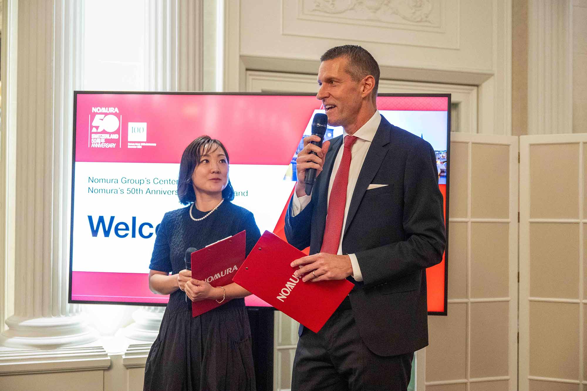 Zwei Personen stehen mit Mikrofonen und roten Ordnern vor einem Bildschirm mit einer Präsentation zur 50-jährigen Jubiläumsfeier von Nomura in einem eleganten Raum.