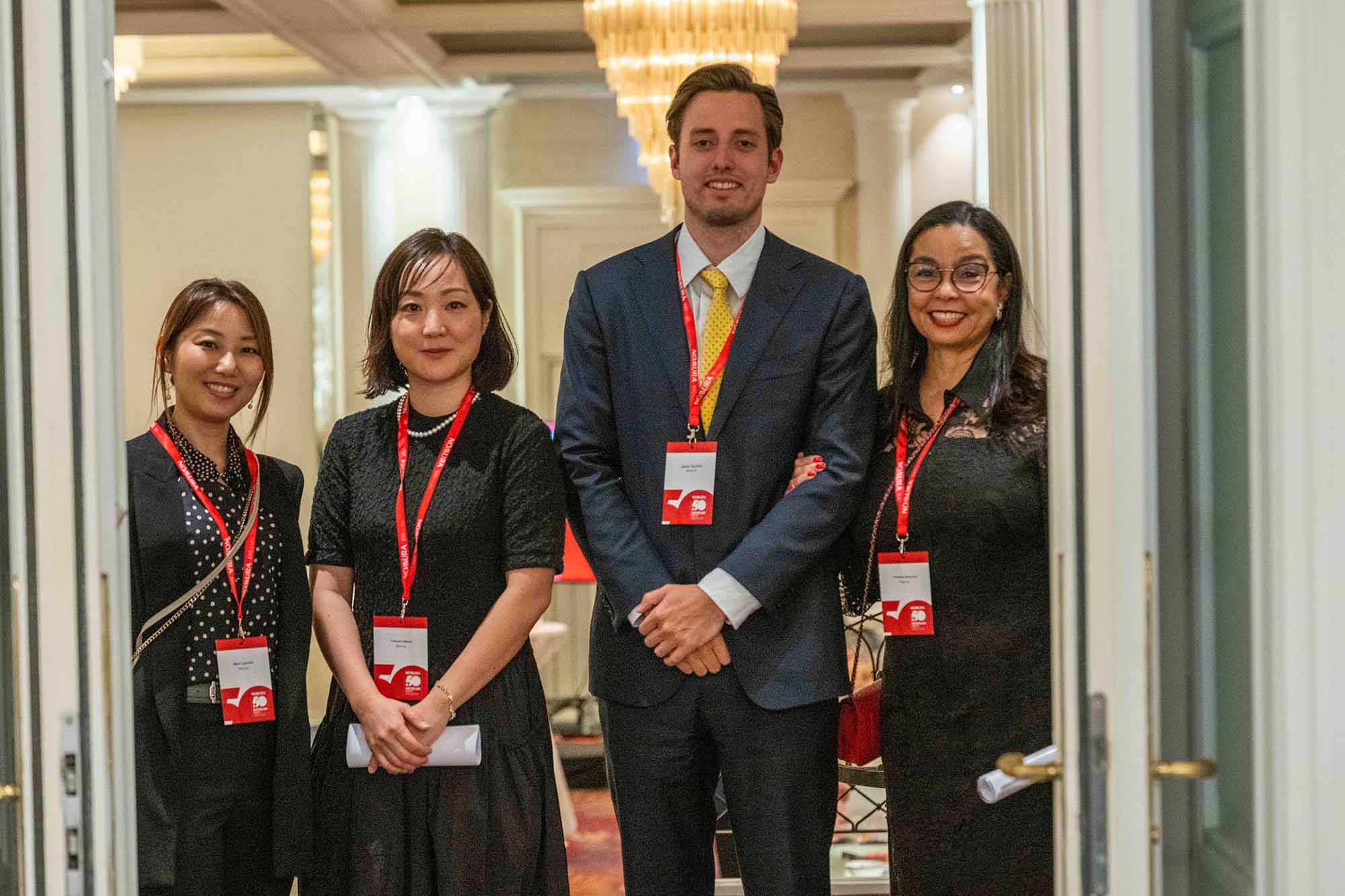 Vier lächelnde Personen mit Namensschildern an roten Bändern stehen in einem eleganten Raum und blicken in die Kamera.