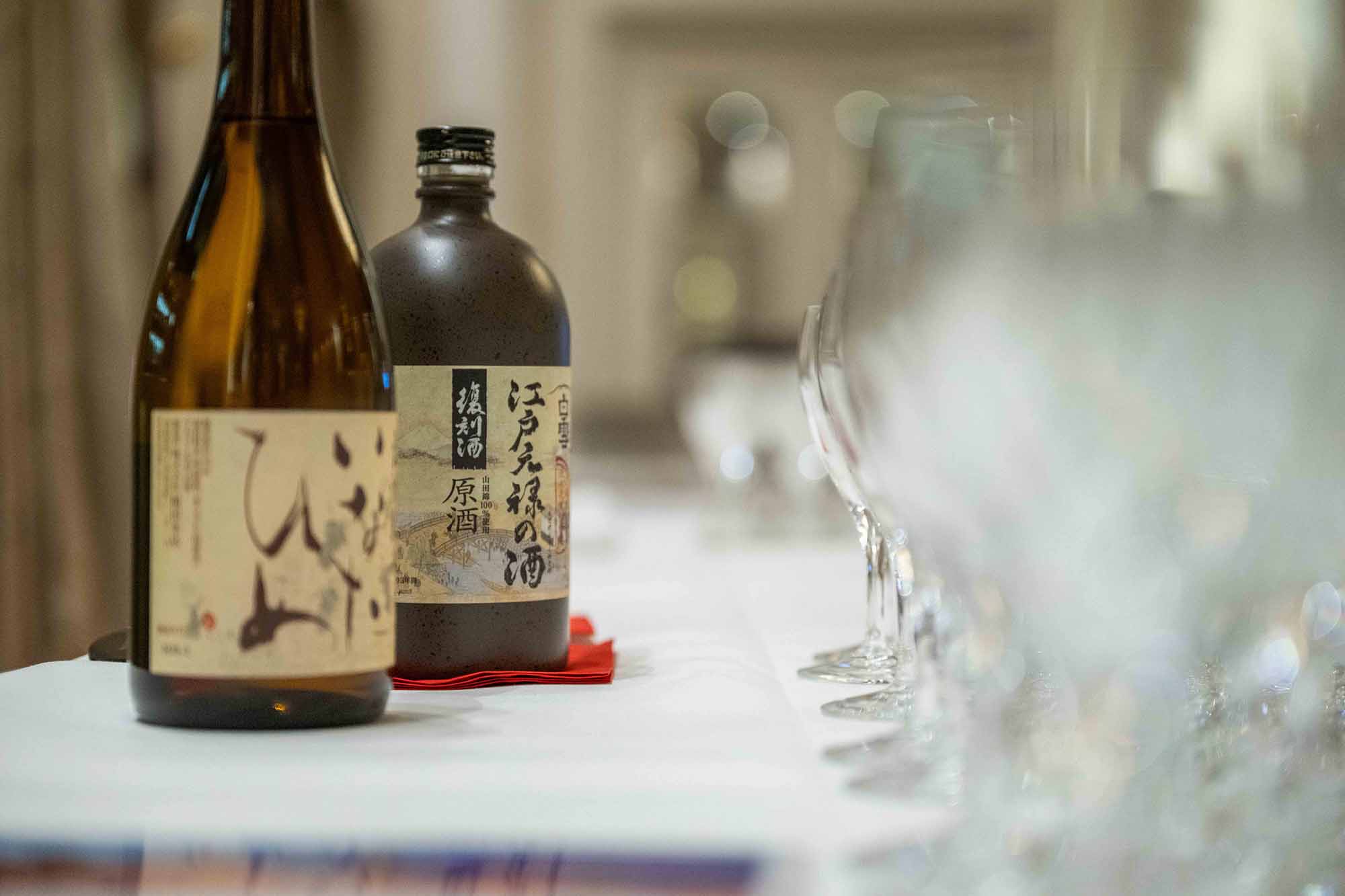 Zwei Flaschen Sake auf einem weißgedeckten Tisch neben einer Reihe leerer Weingläser.