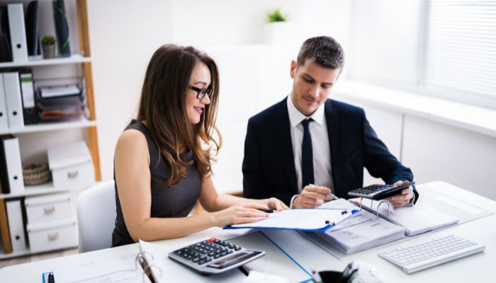 A man and woman in business attire collaborate on paperwork at a desk, focused on their tasks.