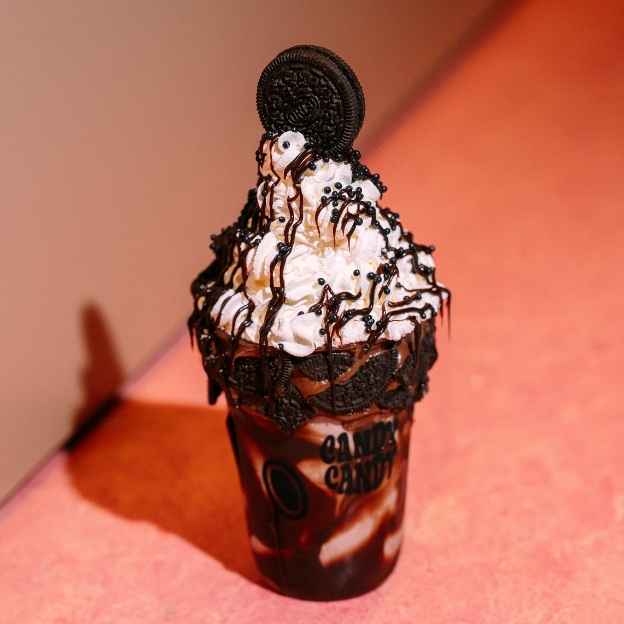 A tall glass filled with a pink milkshake topped with melting vanilla ice cream, colorful sprinkles, and a waffle cone. A wooden spoon rests against the glass.