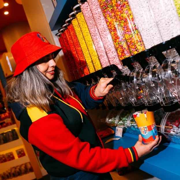 A man in a striped apron operates a crepe maker in a vibrant food stall with colorful decorations in the background. A customer is partially visible on the right.