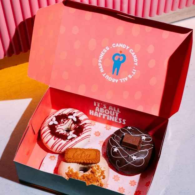 Three pink donuts with colorful sprinkles displayed on a light blue surface, in front of a bright background featuring the words "CANDY CANDY" in playful lettering.