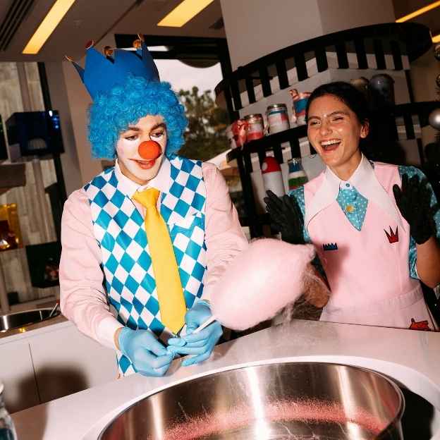 A group of three people wearing colorful outfits and sunglasses, holding clear balloons with 'CANDY CANDY' printed on them. The background features a bright, playful store design.