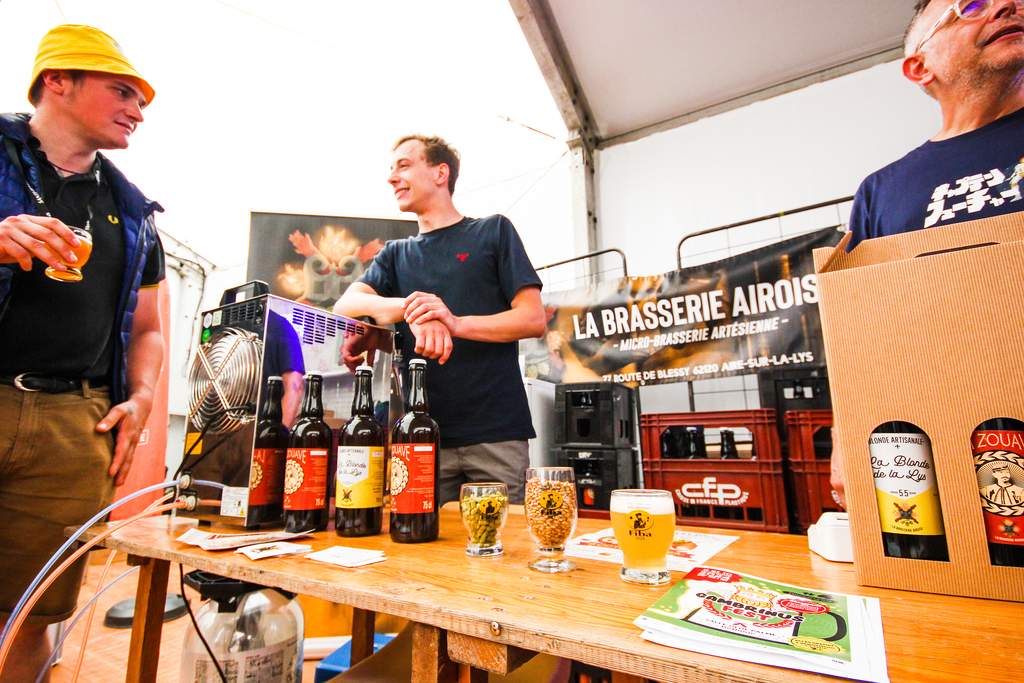Image principale : Un serveur portant un gilet bleu et des bretelles, tenant une planche avec plusieurs grands verres de bière remplis à ras bord. Il se déplace parmi une foule de personnes dans ce qui semble être un festival ou une fête populaire, probablement une fête de la bière. Les personnes autour de lui portent des vêtements traditionnels, et l'ambiance est festive et animée.Miniature : Trois personnes se tiennent près d'un stand de dégustation de bière artisanale. L'une d'elles sert de la bière à partir d'un fût, tandis que des bouteilles de bière et des verres remplis sont disposés sur une table en bois. Une bannière derrière eux indique 'La Brasserie Airois