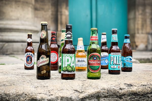 Image principale : Une variété de bouteilles de bière disposées autour d'une caisse en bois décorée pour Noël. La caisse contient des numéros et des décorations festives, suggérant un calendrier de l'Avent ou un cadeau de Noël pour les amateurs de bière.Image secondaire : Une variété de bouteilles de bière posées sur une surface en pierre devant un fond de porte turquoise. Les bouteilles représentent différentes marques et types de bière du monde entier.