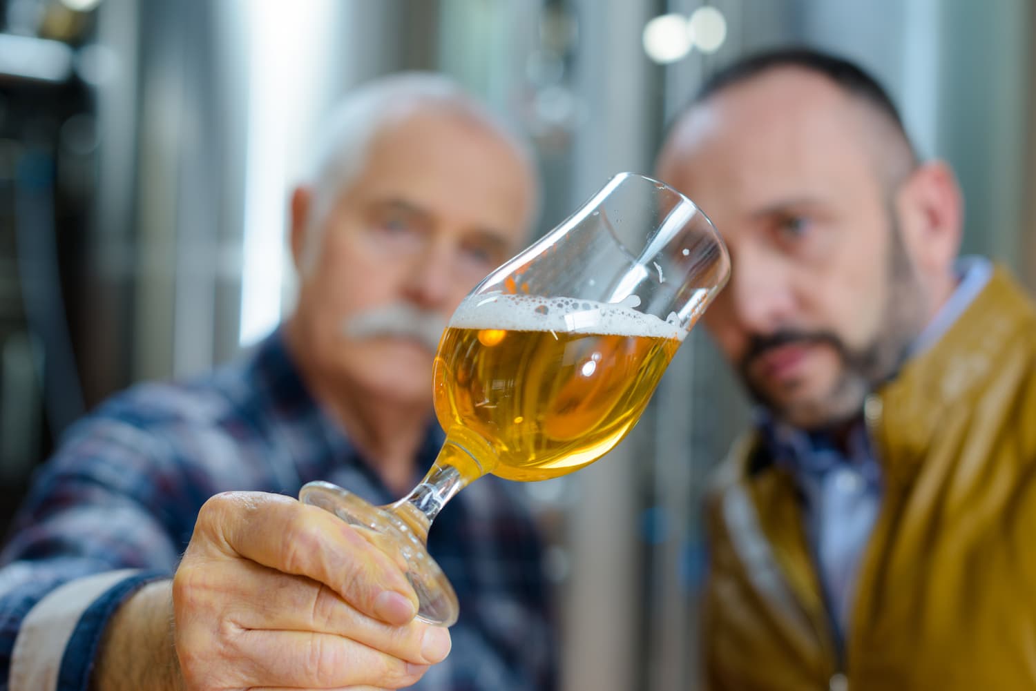 image principale : "Un homme souriant avec des lunettes et une casquette, remuant un mélange dans un grand bol, entouré d'équipements de brasserie et de décorations de Noël. Il semble préparer une recette spéciale ou expérimenter de nouvelles saveurs, dans une ambiance festive et créative. image miniature : Deux hommes dans une brasserie, l'un tenant un verre de bière à la lumière pour examiner sa couleur et sa clarté. L'arrière-plan montre des équipements de brassage en acier inoxydable.