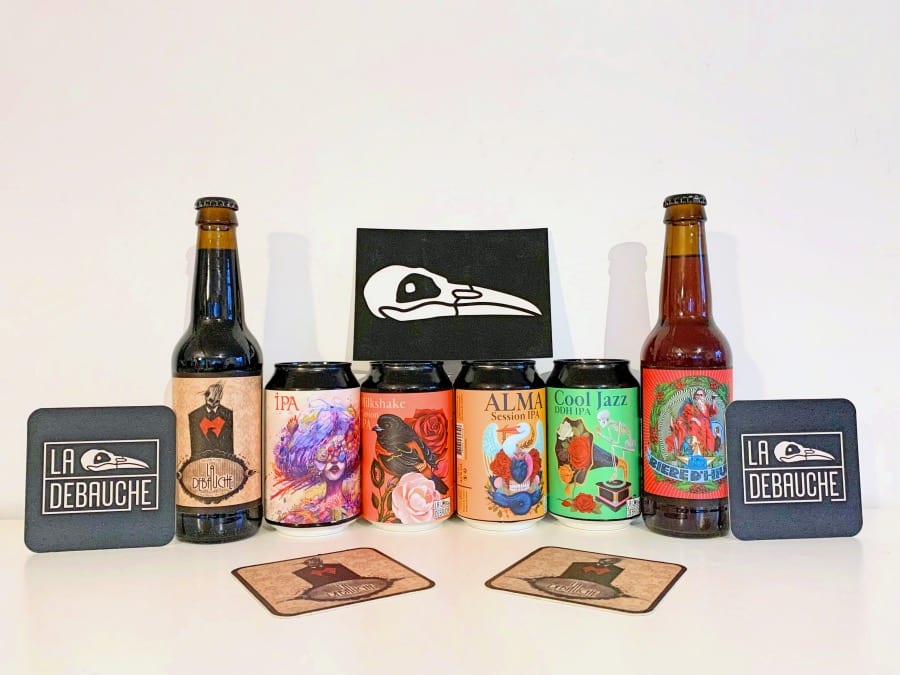 Image principale : Une sélection de bouteilles de bière avec des étiquettes colorées, posées devant des cuves de brasserie avec un homme debout derrière. L'homme semble être un brasseur ou un employé de la brasserie, présentant fièrement les différentes variétés de bière produites."Miniature : Une sélection de bières et de sous-verres de la marque 'La Débauche'. Les bouteilles et canettes présentent différentes variétés de bière, dont une IPA, une Double IPA, et une bière de Noël. Les sous-verres portent le logo de la marque.