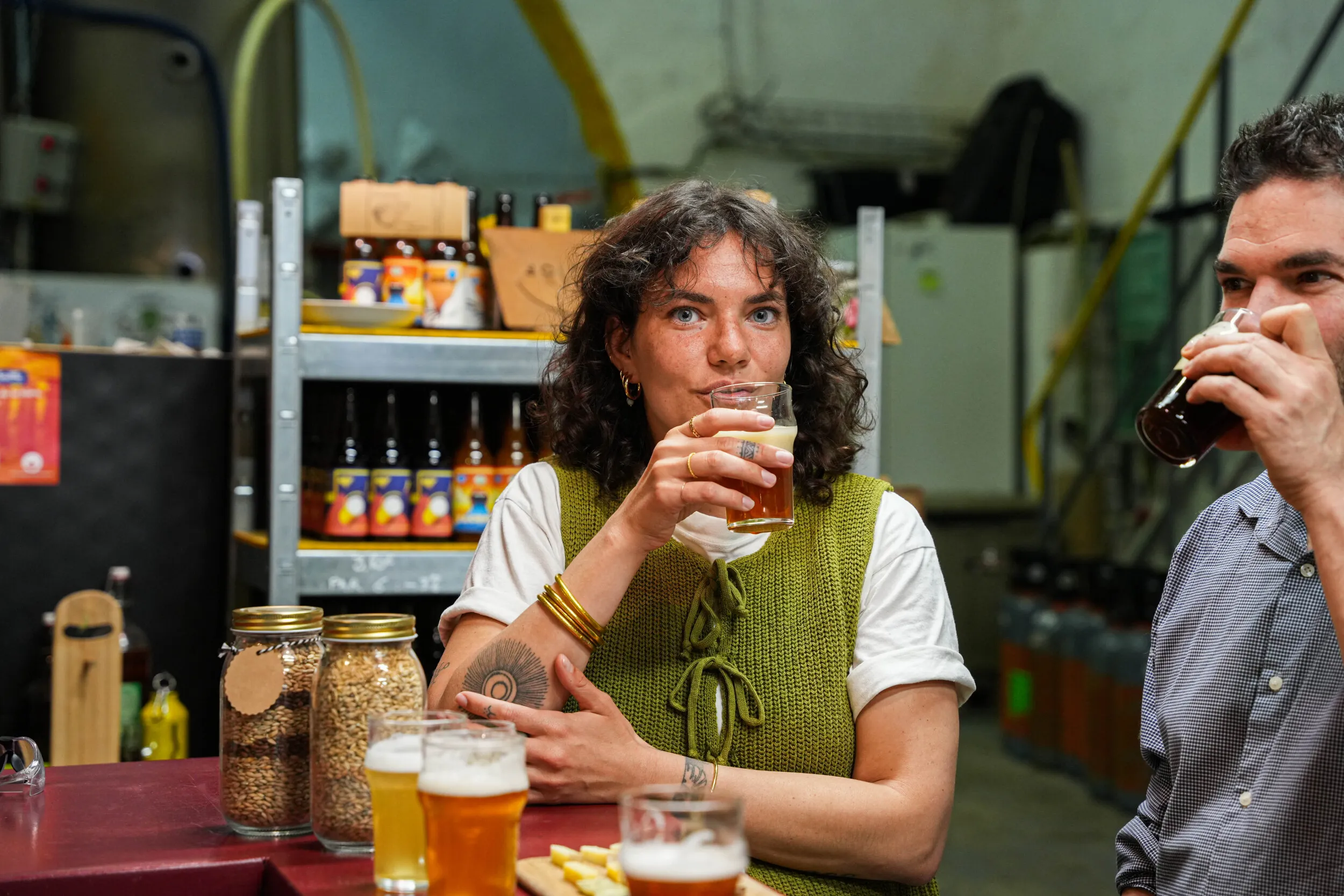 Image Principale : Un homme en train de sentir des verres de bière de différentes couleurs, alignés sur une table devant lui. Il semble évaluer les arômes des différentes bières, peut-être dans le cadre d'une dégustation ou d'une évaluation sensorielle." Miniature : Deux personnes assis à une table dans une brasserie, dégustant de la bière. La femme porte un gilet vert et tient un verre de bière, tandis que l'homme à côté d'elle boit également de la bière. Sur la table, il y a des verres de bière et des bocaux de grains.