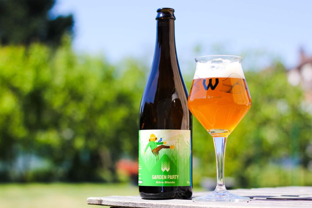 Verre de bière artisanale French Farmhouse avec un logo distinctif en forme de 'W', accompagné d'une bouteille de bière en arrière-plan, illustrant l'ambiance conviviale de L'instant bière.