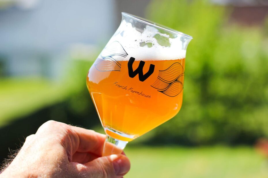 Verre de bière artisanale French Farmhouse avec un logo distinctif en forme de 'W', accompagné d'une bouteille de bière en arrière-plan, illustrant l'ambiance conviviale de L'instant bière.