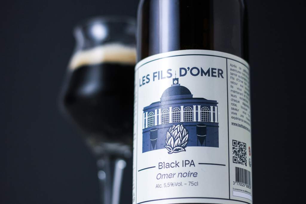 Bouteille de bière artisanale 'Les Fils d'Omer' de type Black IPA, avec une étiquette illustrée d'un bâtiment classique et un verre de bière noire en arrière-plan, mettant en valeur la diversité des saveurs proposées par L'instant bière.