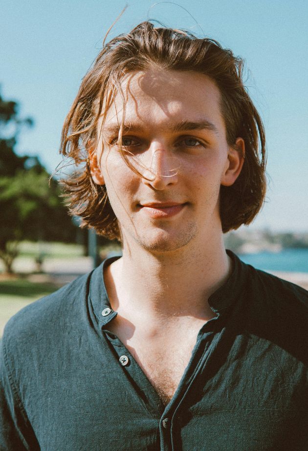Un homme aux cheveux longs, souriant, avec un tshirt vert foncé se tient devant un plan d'eau calme.