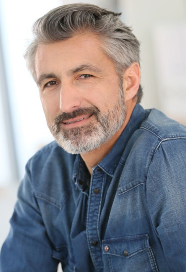 Un homme souriant avec une barbe grisonnante, portant une chemise en jean bleu, se tient dans un environnement intérieur lumineux