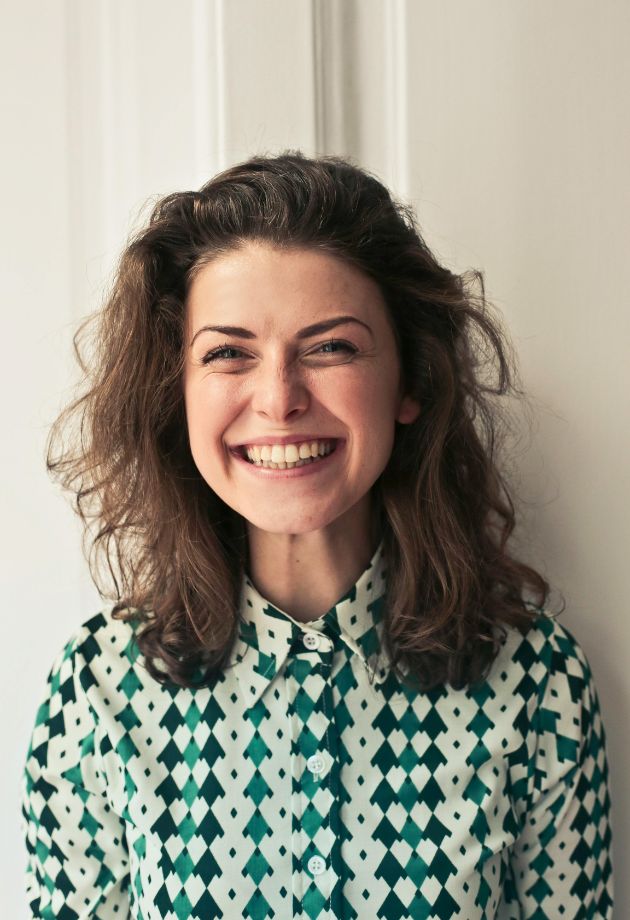 Une femme souriante avec des cheveux bruns ondulés, portant une chemise à motifs géométriques verts et blancs, se tient devant un fond blanc.