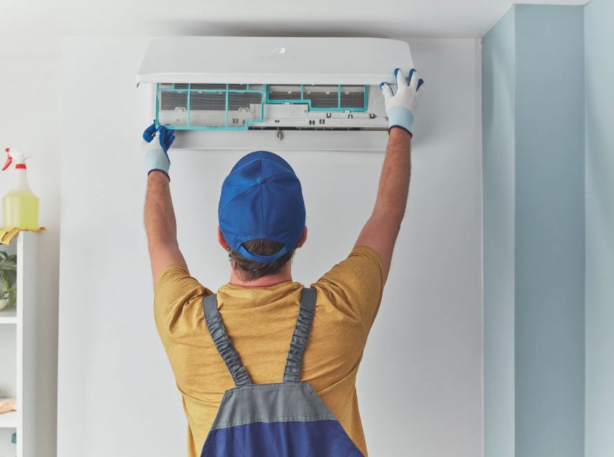 HVAC technician wearing a blue cap and gloves servicing a wall-mounted air conditioning unit in a home.