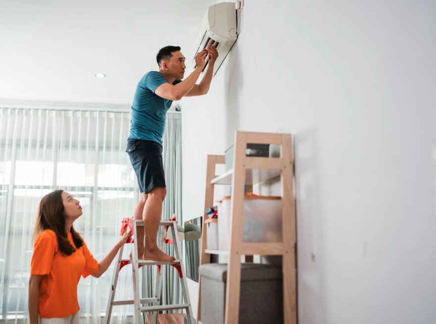 Man standing on a ladder repairing a wall-mounted air conditioner while a woman looks on in a modern living room.