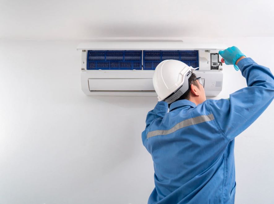 Technician wearing a hard hat and blue uniform repairing a wall-mounted air conditioning unit with a screwdriver.