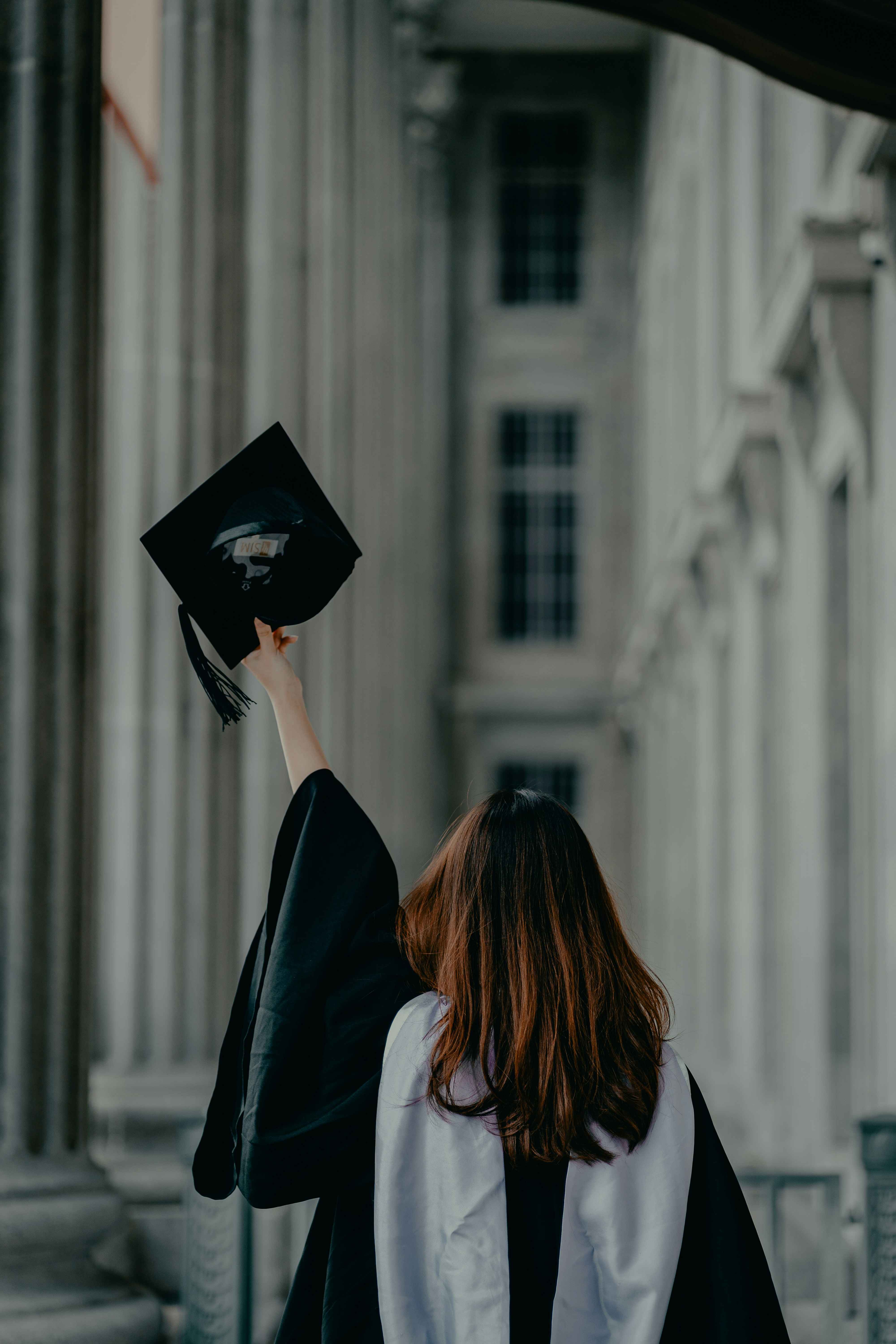 A woman photographed from behind on her graduation day, raising her left arm and holding her graduation cap in her hand, celebrating her achievement.