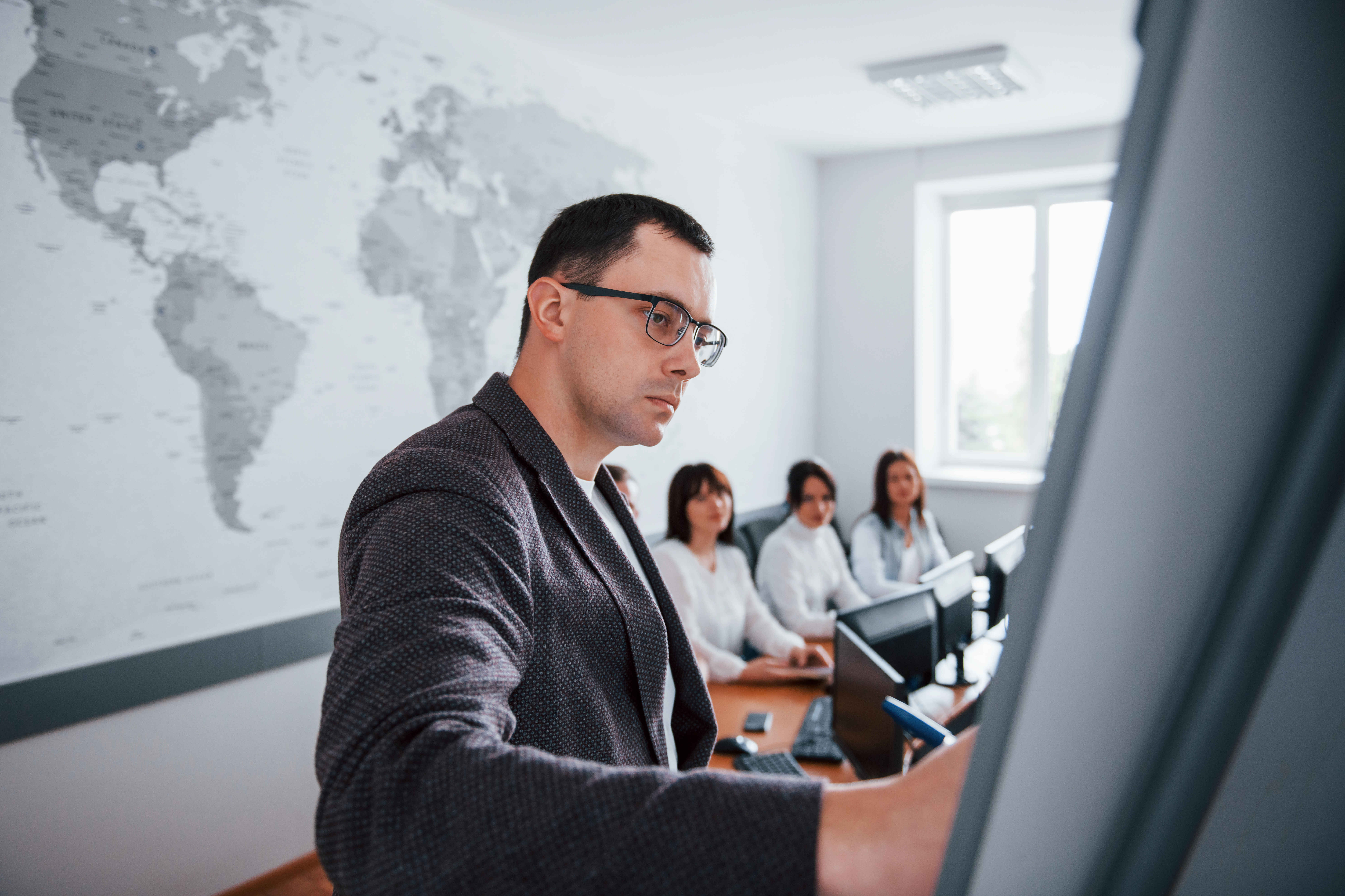 A serious-looking lecturer is standing in a computer lab classroom, pointing to or showing something on a board to a small group of students gathered around, who are paying close attention.