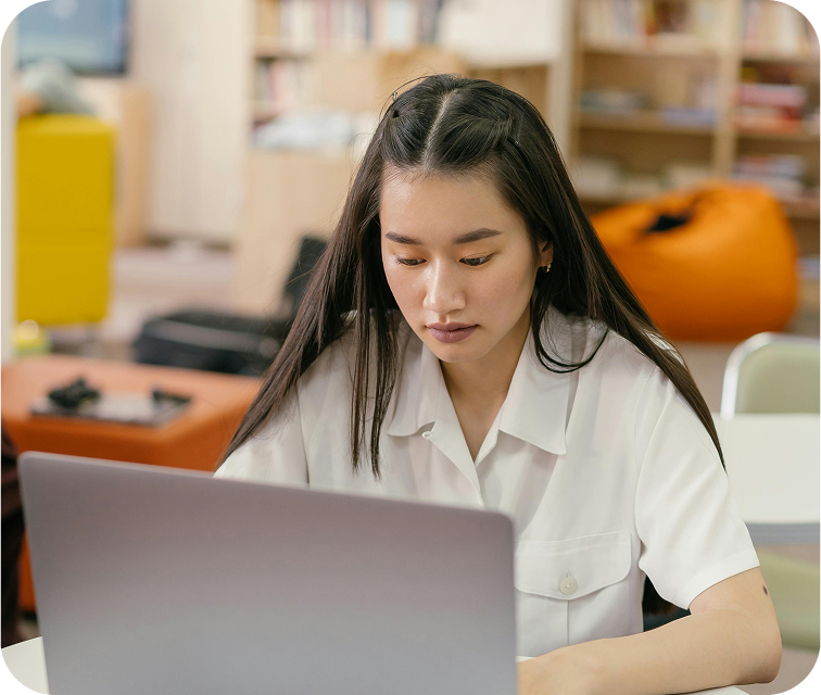 A young Asian female student is seated, looking serious and focused as she concentrates on the screen of her laptop.