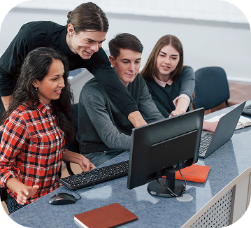 A group of four students are gathered around a desktop monitor, appearing focused and engaged as they study together.