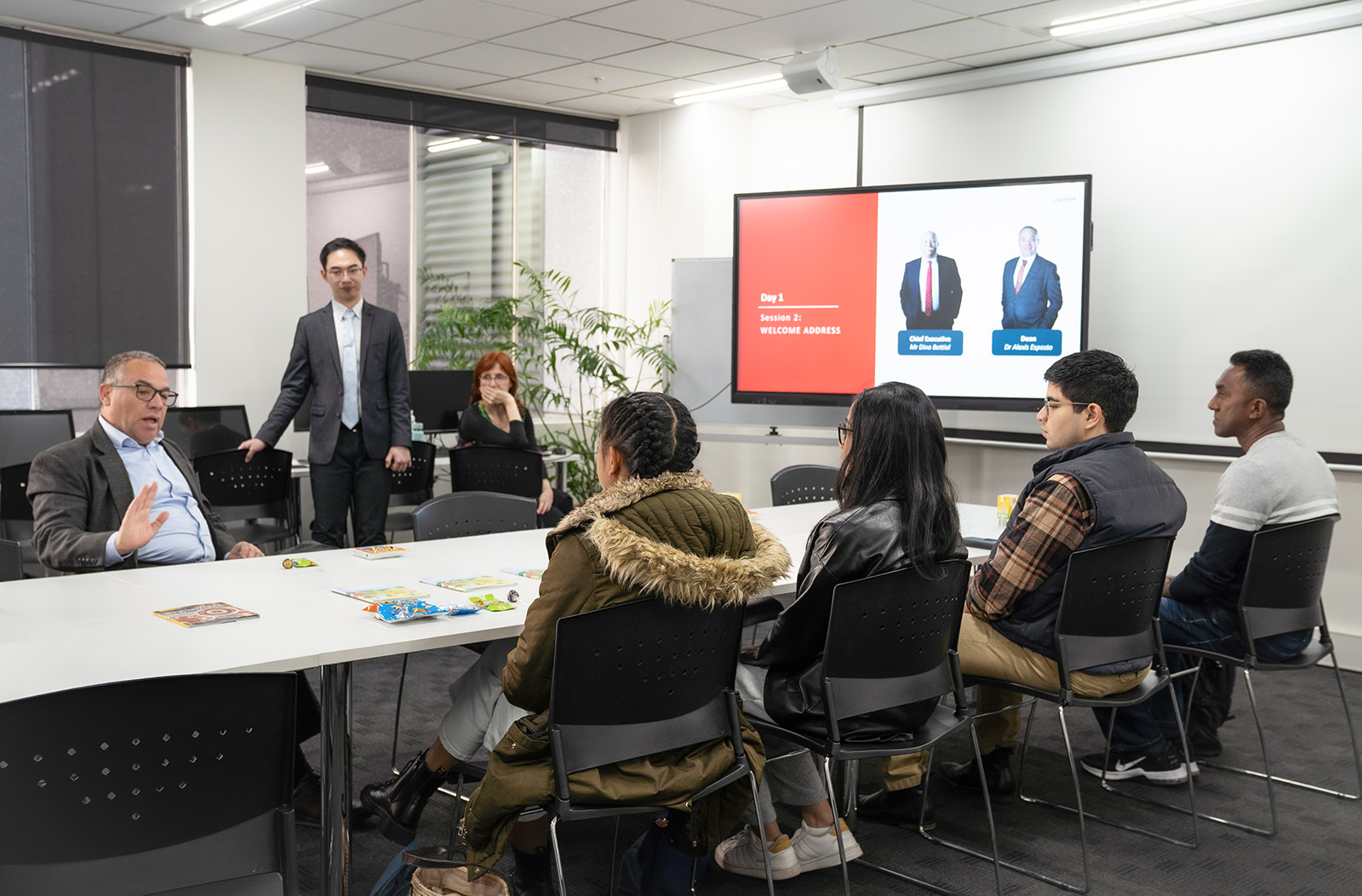 A group of students is seated around a large rectangular table, attentively listening to three professors or lecturers during an orientation session at Danford Higher Education library.