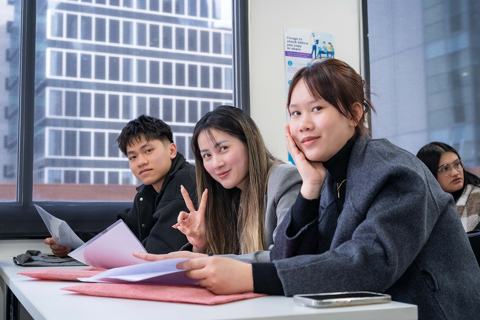 Three students are seated together during a class session, smiling warmly at the camera. One student, a young woman, holds up her hand making a victory sign, adding a sense of playfulness and camaraderie to the moment.