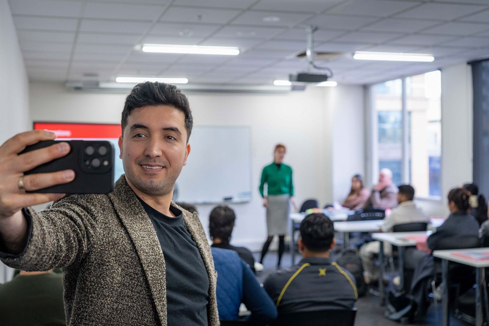 A lecturer is taking a selfie in a classroom. In the background, a class session is visible but blurred, with students seated and engaged.