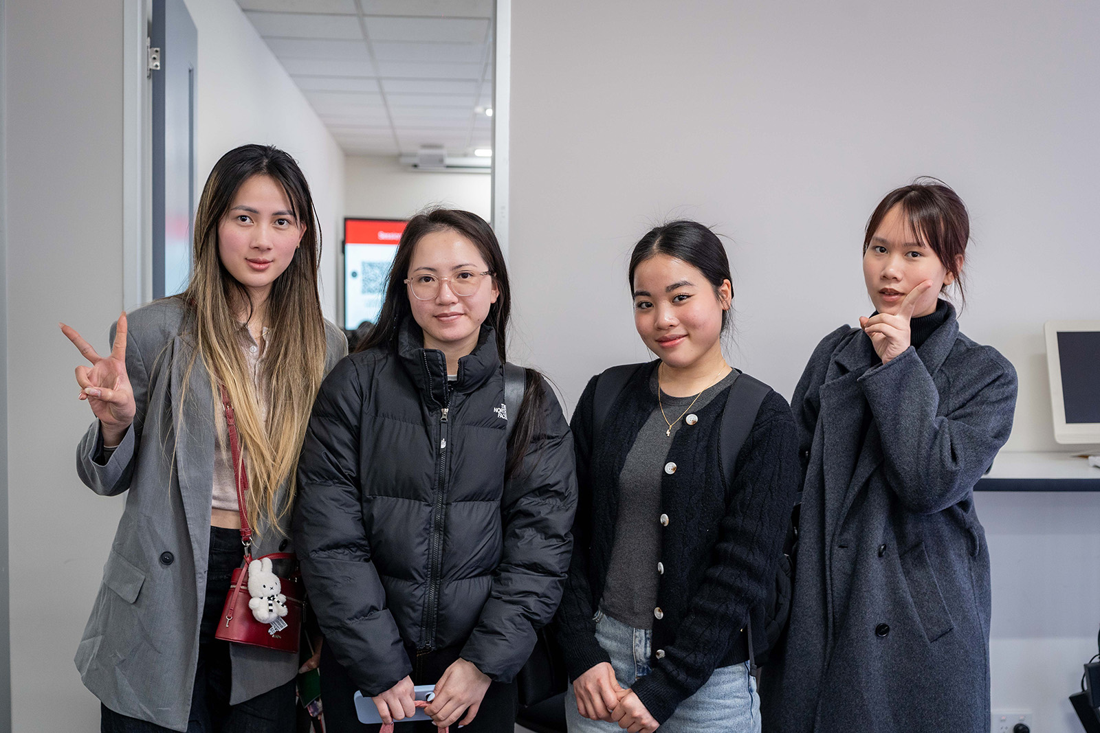 Four young female students are standing in front of a classroom door, posing for the camera with gentle smiles. Their relaxed expressions convey a friendly, welcoming atmosphere.