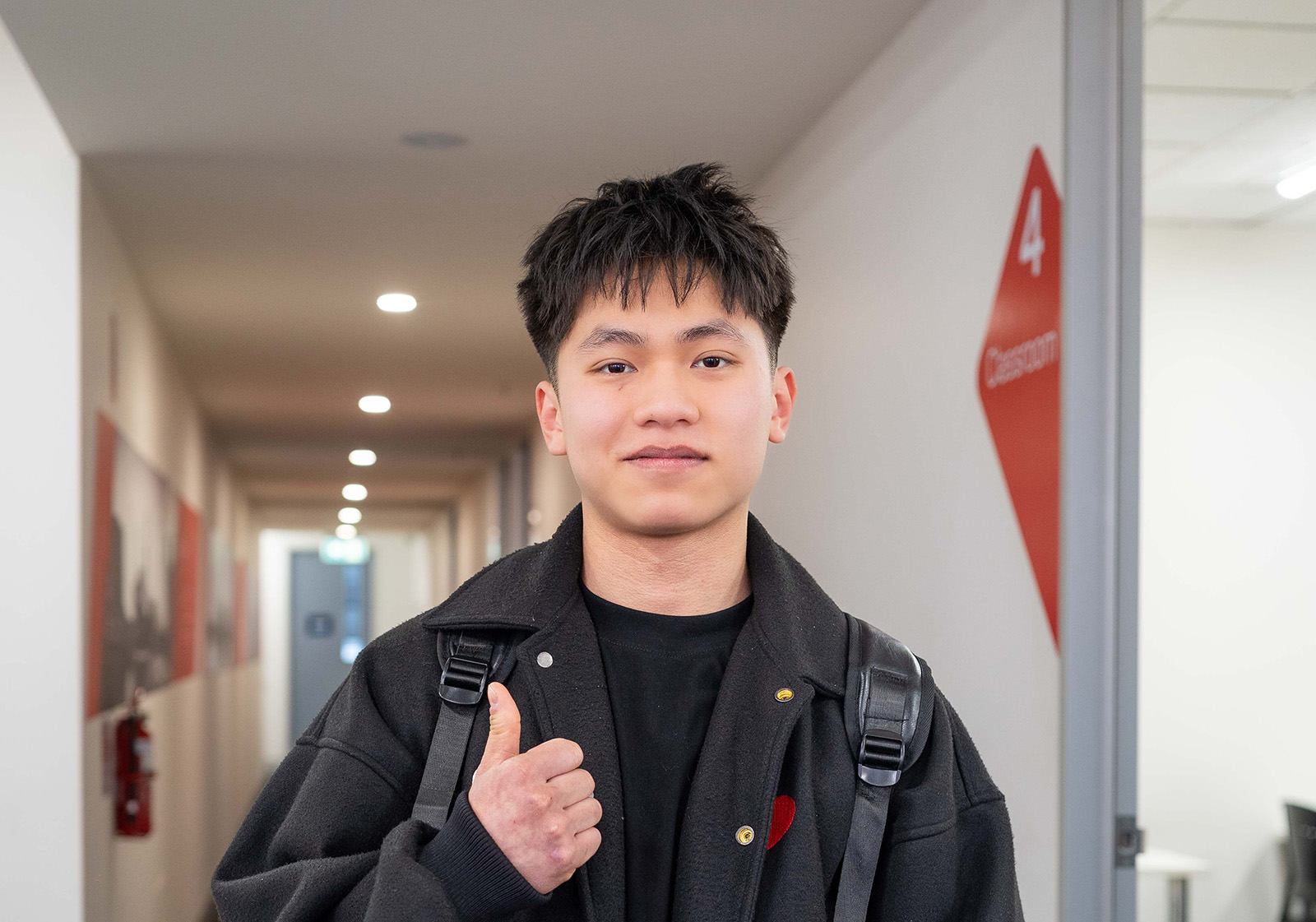 A young male student of Asian background is smiling and giving a thumbs up to the camera. He represents international students and conveys a positive, welcoming attitude.