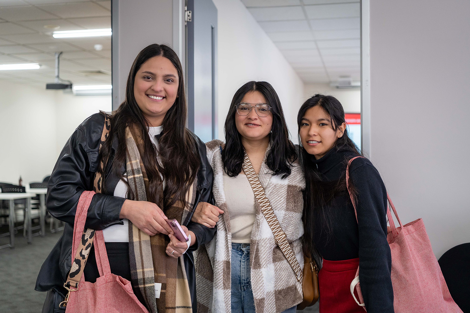 Three young female students are posing for the camera, smiling. Behind them, a hallway of the campus is visible.