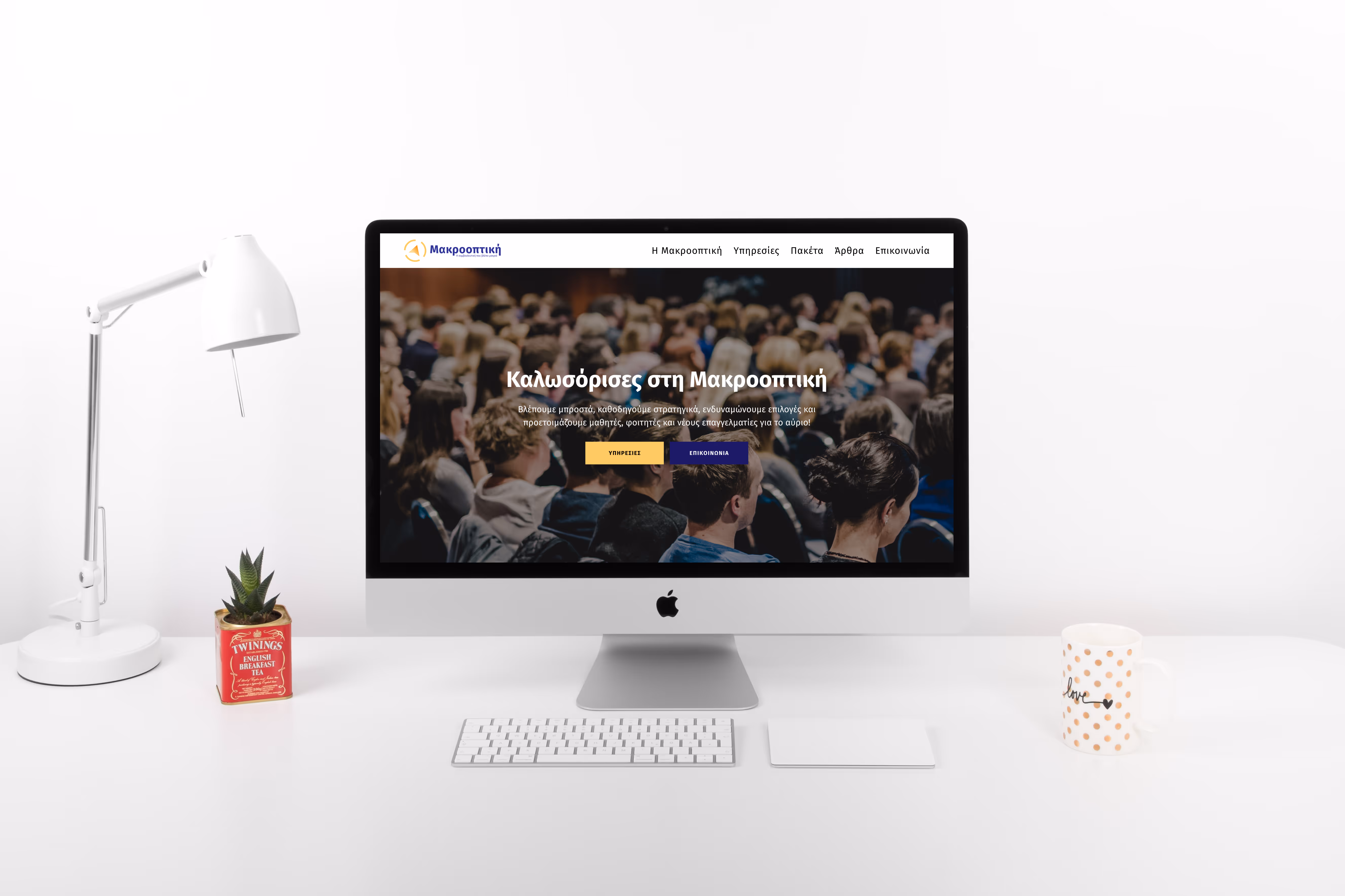 iMac desktop on white desk displaying a website in Greek with a crowd background, next to a white lamp, small plant in red Twinings tea tin, and a white mug with orange polka dots.