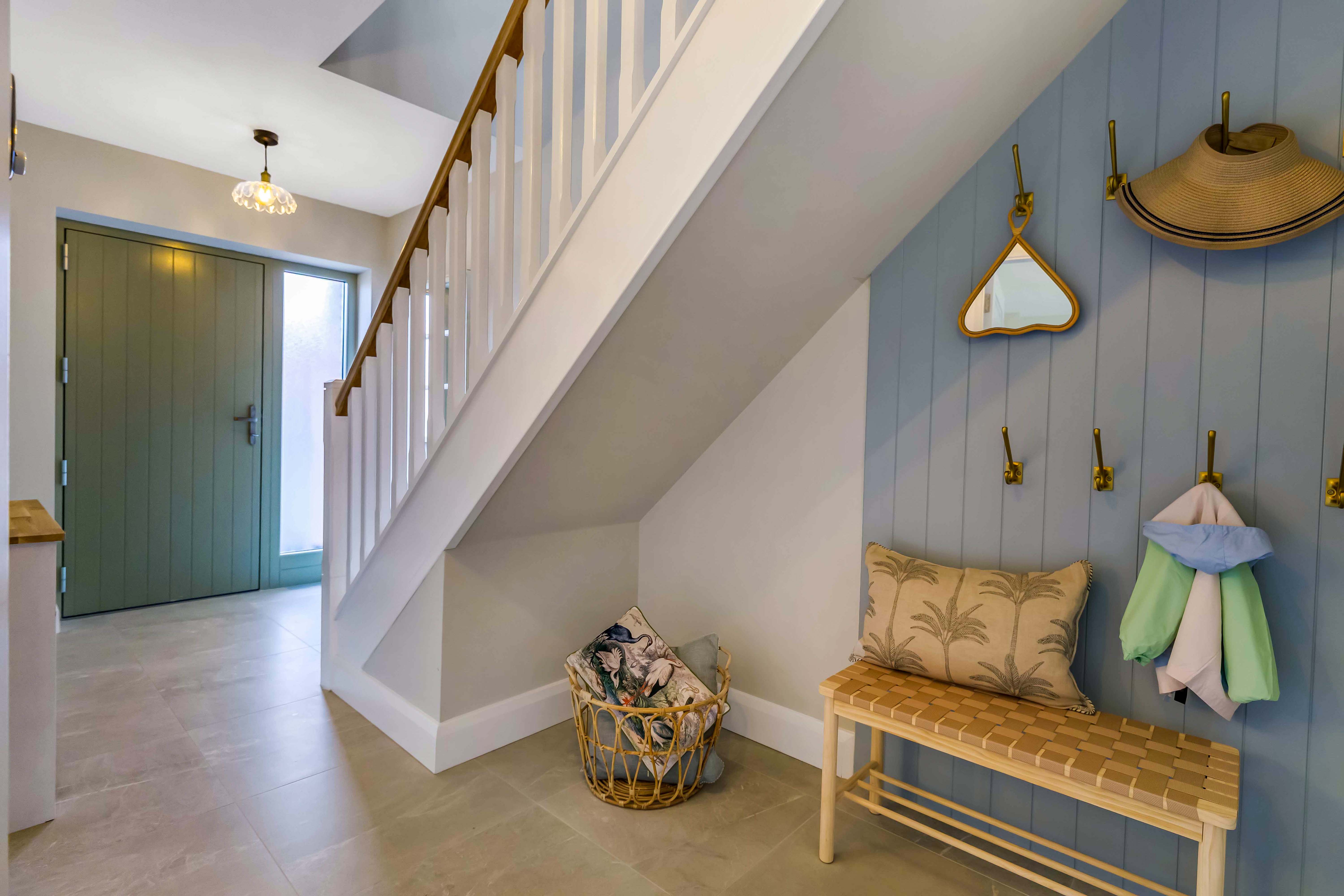 Entryway with light green door, white staircase, basket with pillows, bench, and blue paneled wall with hooks.