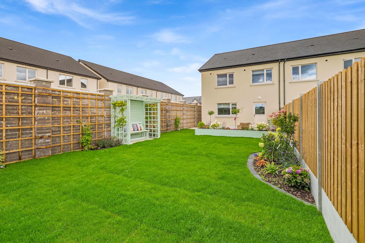 Green backyard with a wooden bench pergola, flower beds, and fenced yards around homes.