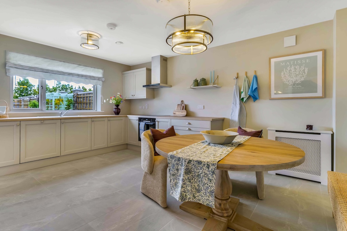 Modern kitchen with beige cabinets, round wooden dining table, pendant lights, and a window with garden view.
