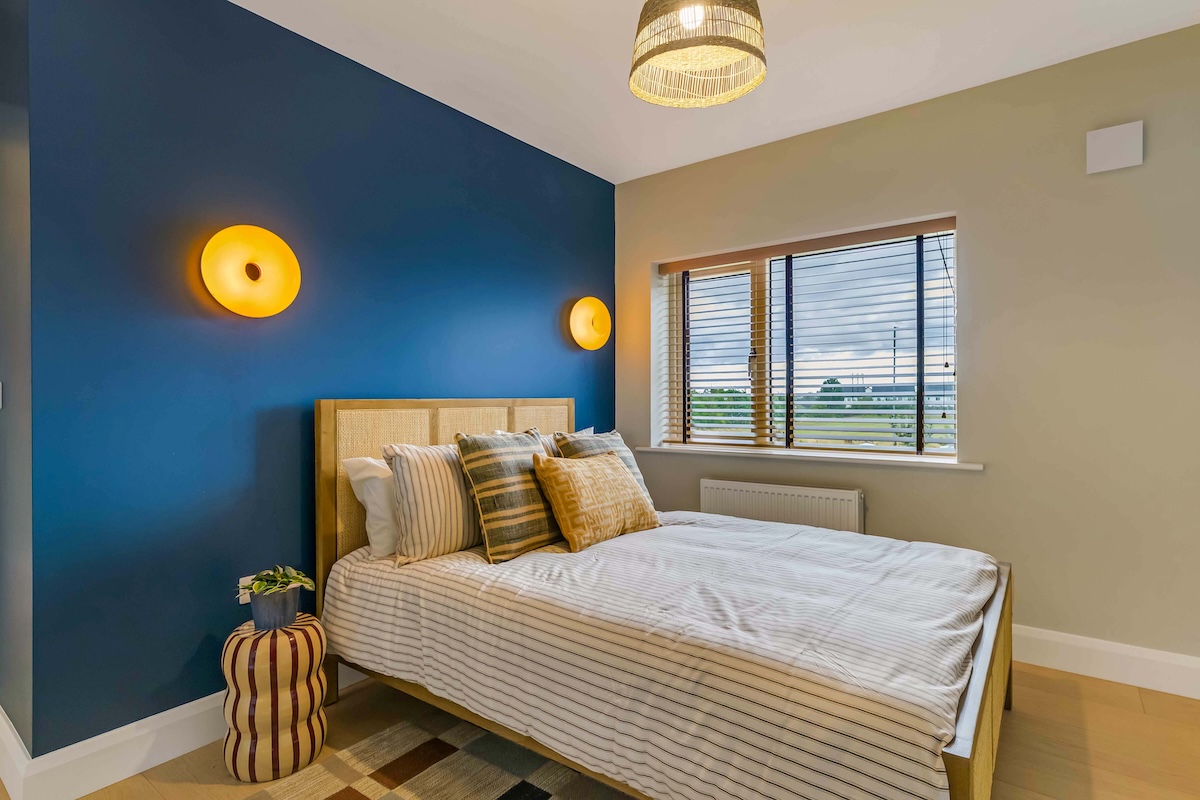 Bedroom with wooden bed, striped bedding, blue accent wall, round yellow wall lights, and window blinds.