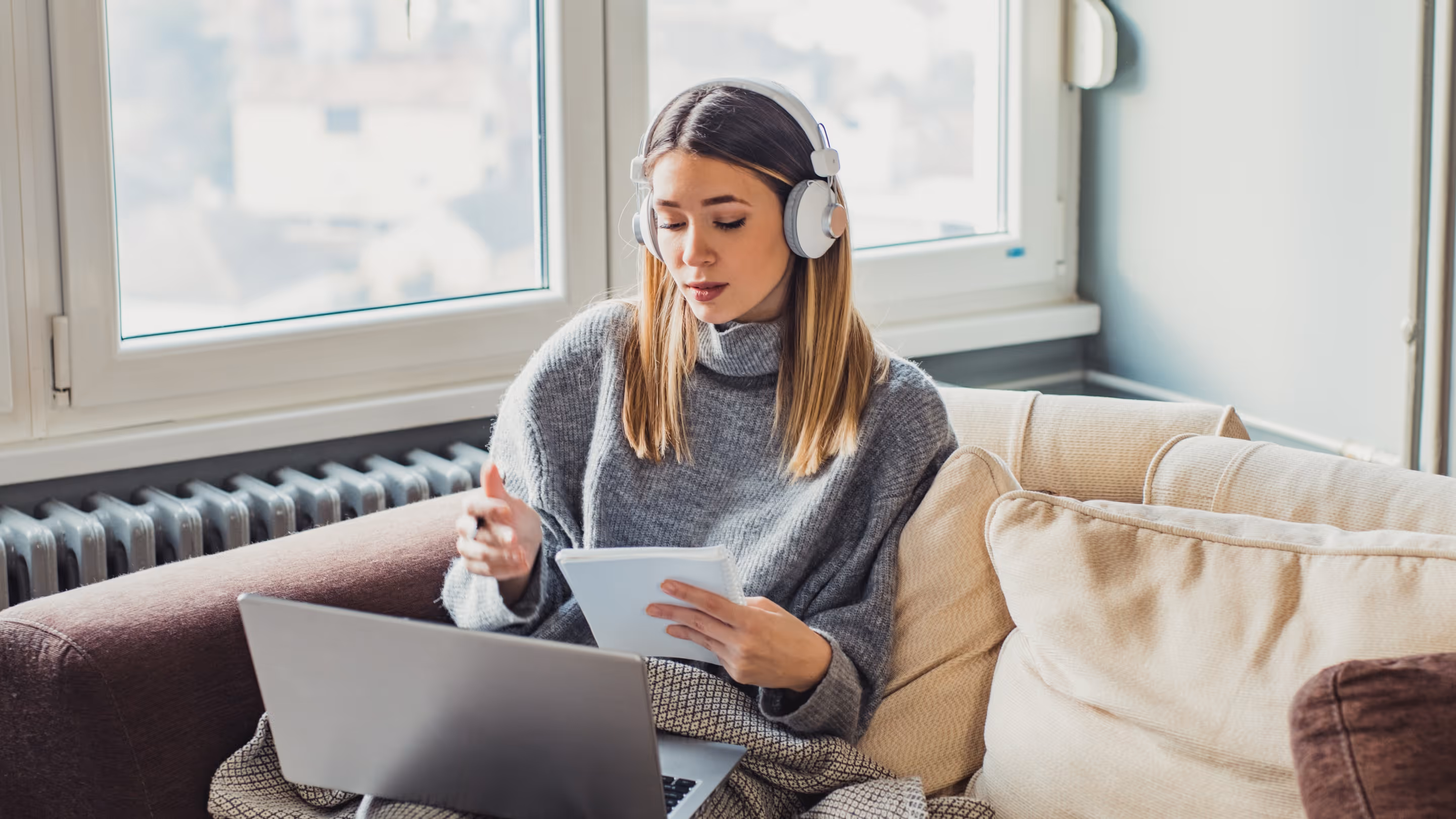 Woman with headphones sitting on a couch using a laptop and holding a notepad in a bright room.