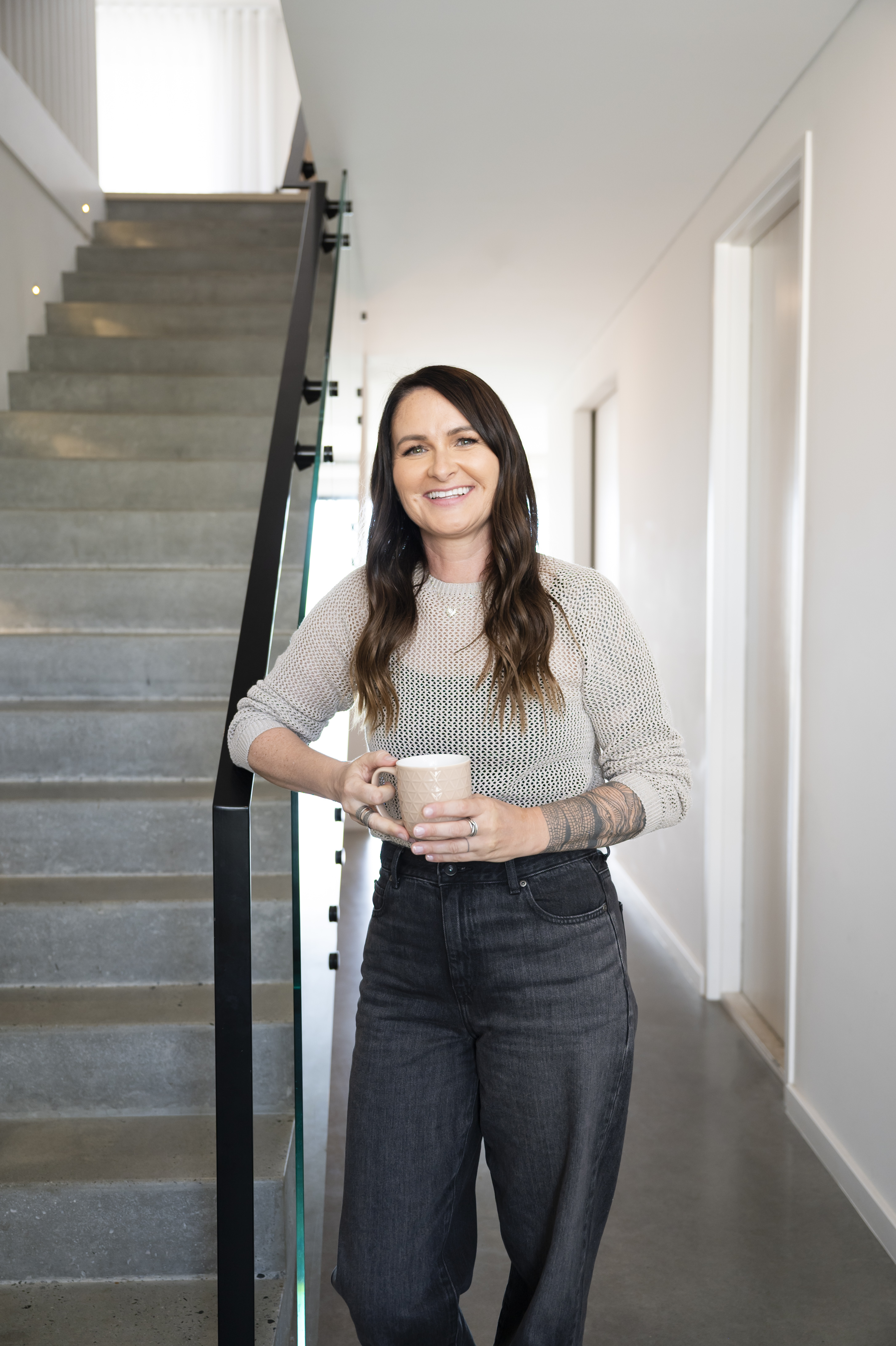 Smiling woman in brown blazer and jeans leaning on a wooden dining table in a bright kitchen.