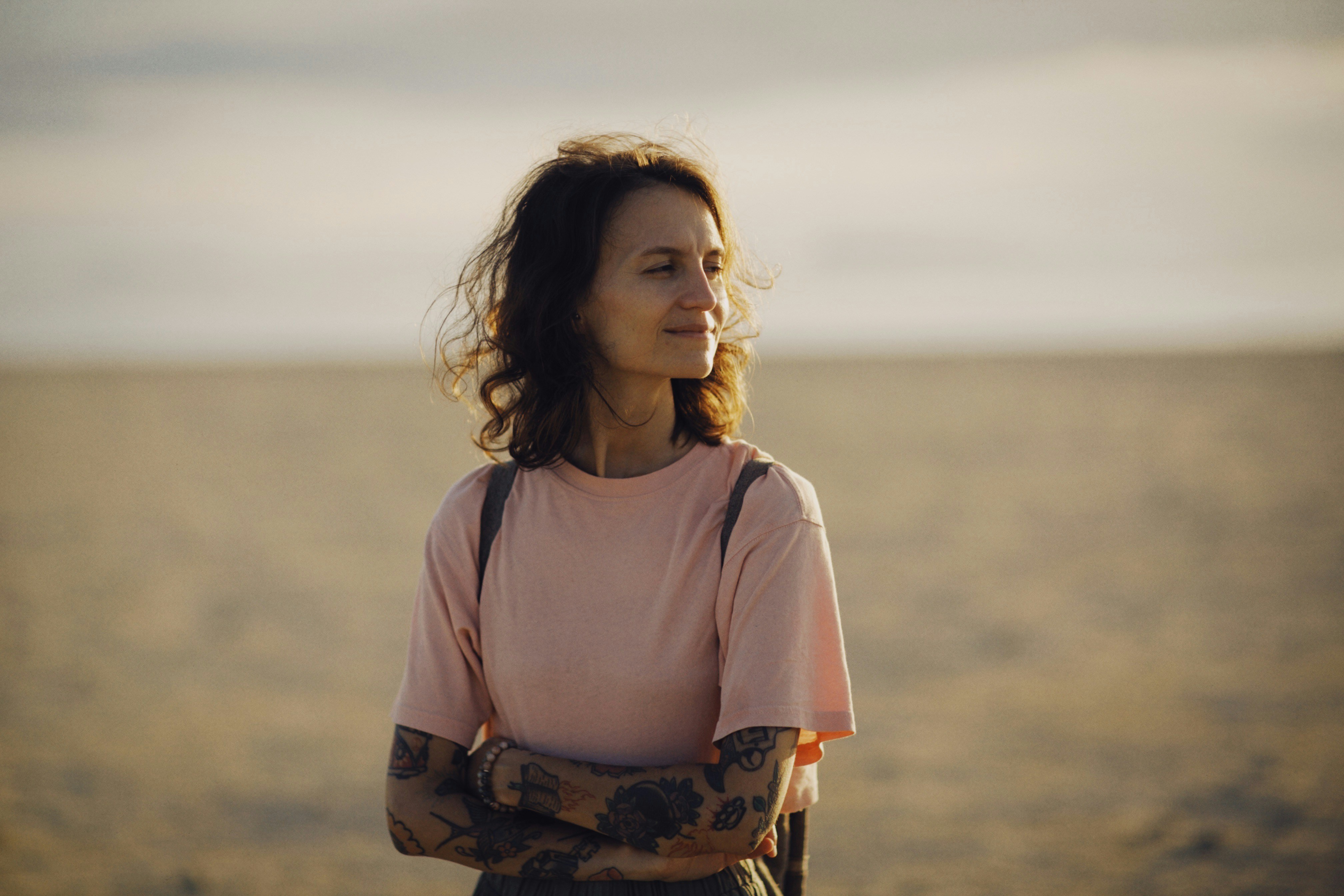Woman with curly hair wearing a pink t-shirt and tattoos on her arms stands on a sandy beach with arms crossed, looking to the side with a slight smile.